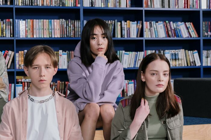 Three solemn teens, looking at the camera, with a bookshelf background, feeling second-hand embarrassment from a cringe situation.