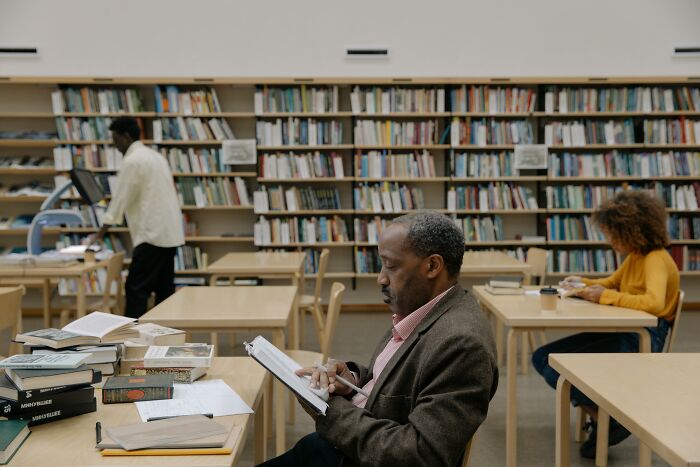 A man studies literature in a library, surrounded by bookshelves. Others also read and study, proving their smarts.