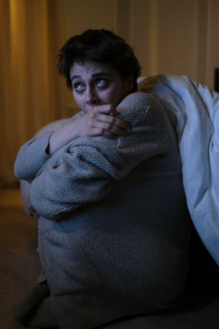 A scared woman in a dark room, hugging her knees, looking up anxiously. She just experienced creepy things.
