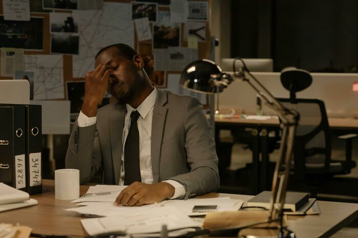 A lawyer in a gray suit looks stressed at his desk, head in hand, surrounded by case files. Hardest cases.