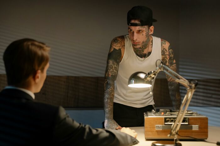A man covered in tattoos, wearing a tank top and backward cap, intimidating a man in a suit during a hard case.