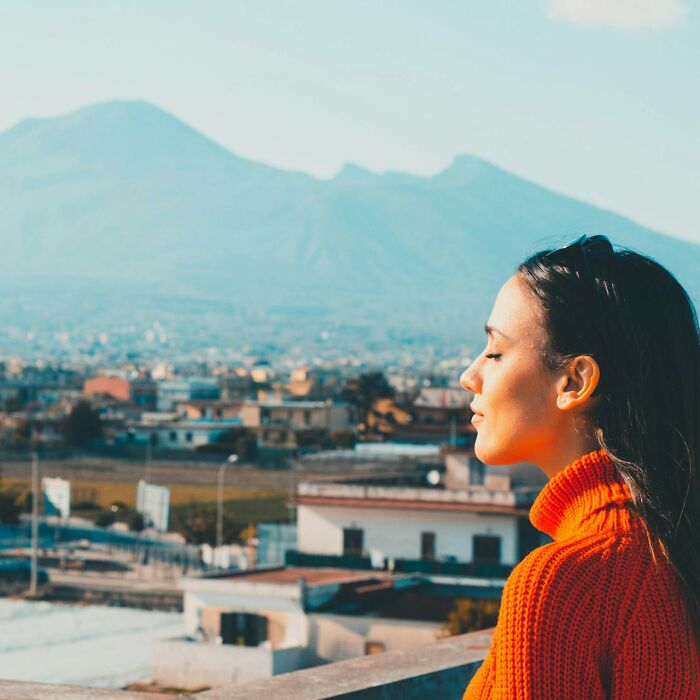 A woman in an orange sweater, eyes closed, enjoying a sunny view of a city and mountains. #SilentLuxuries #RichPeople
