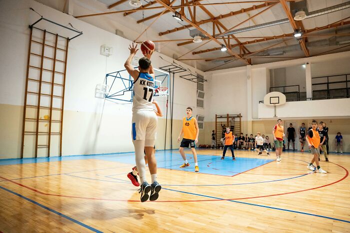 A young basketball player in mid-air, shooting the ball. Other players and spectators are blurred in the background, possibly experiencing second-hand embarrassment.