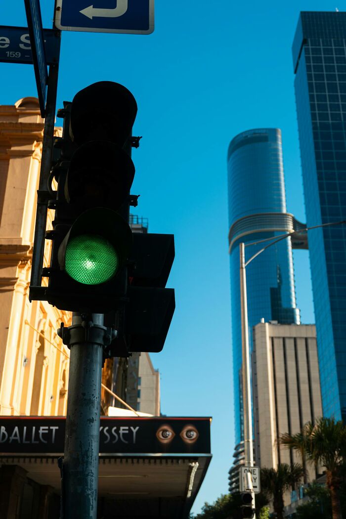 A street scene with a glowing green traffic light. Tall buildings and a blue sky are visible in the background, a glitch in the matrix.