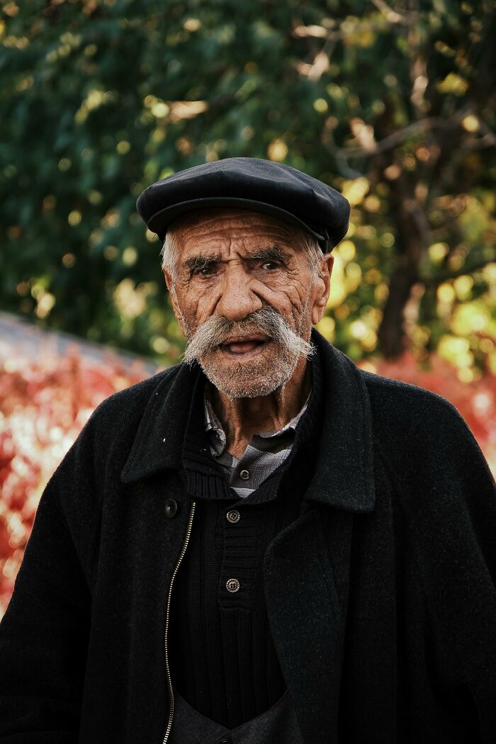 Older man with mustache and hat standing outside with leafy background