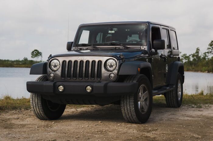 Dark grey Jeep Wrangler parked on a dirt path beside a lake, illustrating Glitches In The Matrix themes.