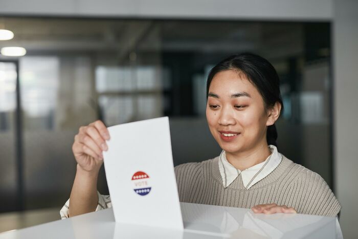 A young Asian woman, a Gen Z, smiles as she inserts a ballot with a "VOTE" sticker into a box.