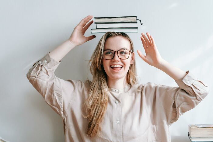 A happy woman in glasses balances books on her head, excited to find out if her IQ is above average with this quiz.