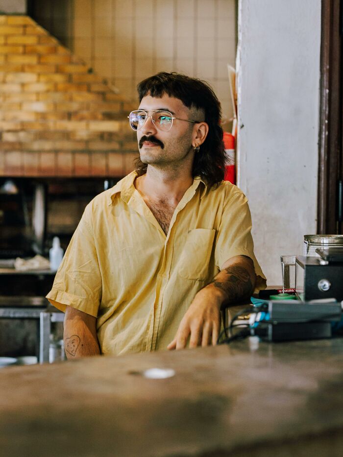 A Gen Z man with a mustache, mullet, and glasses, wearing a yellow shirt, leaning on a counter, embodying unique generational trends.