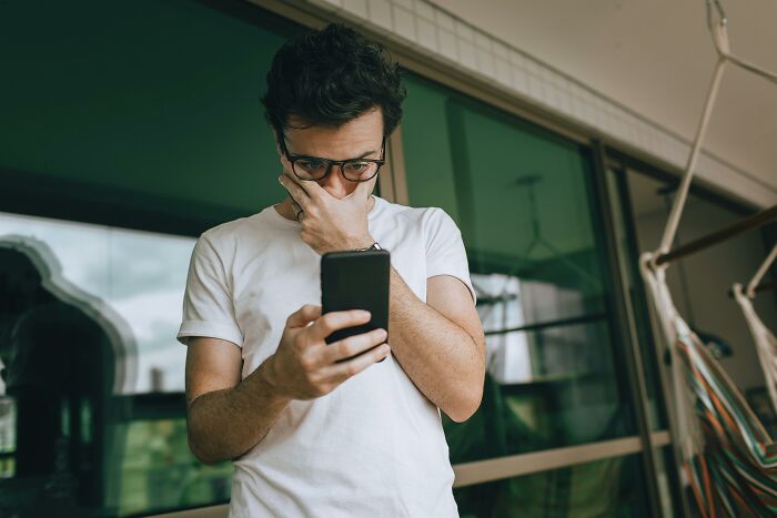 A man in glasses looks shocked at his phone, hand covering his mouth, suggesting he found out his partner was cheating.