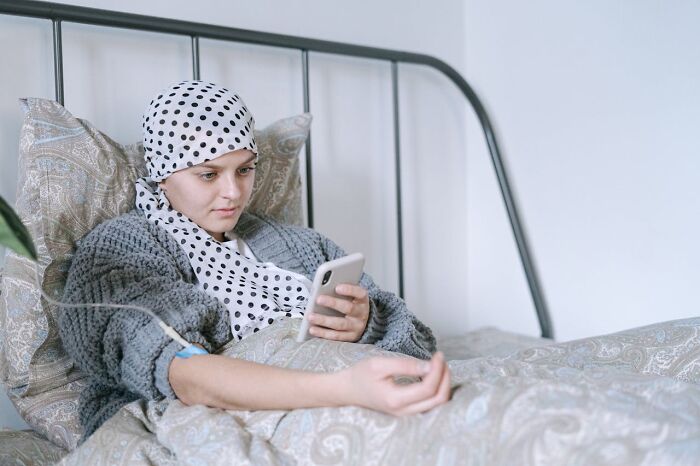 A woman with a polka dot headscarf and an IV in her arm lies in bed, looking at her phone. An example of deeply immoral things.