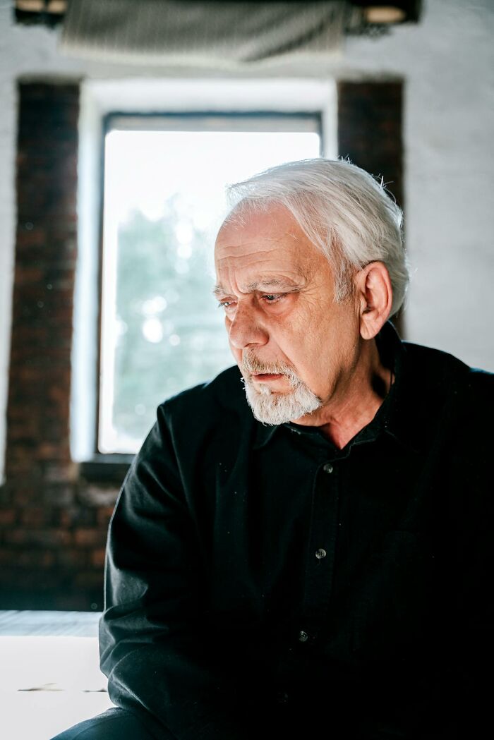 An older man with gray hair and a goatee, wearing a black shirt, looks down thoughtfully, reflecting on deeply immoral things.
