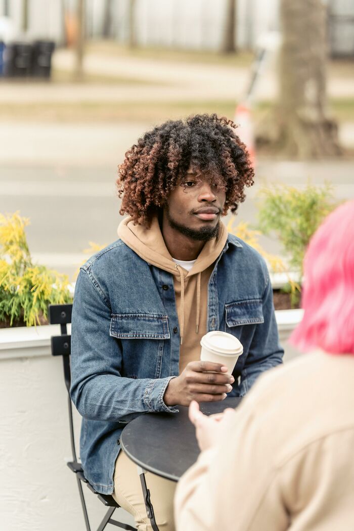A Gen Z man with curly hair and a denim jacket, holding coffee, listening to a pink-haired person.