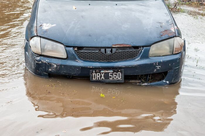 A submerged blue car in muddy floodwaters. The damaged vehicle is half-submerged, reflecting the disturbing d***h stories of natural disasters.