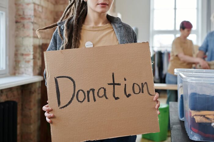 A person with dreadlocks holds a cardboard sign with "Donation" written on it, highlighting deeply immoral actions and kindness.