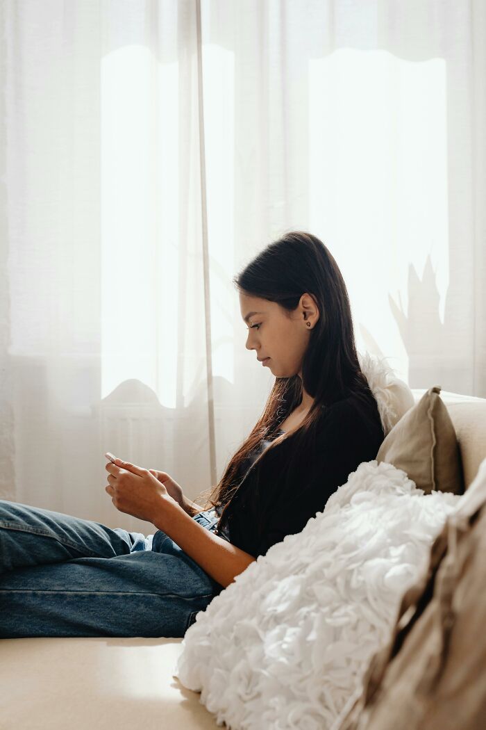 Young woman on a sofa, looking at her phone. She is wearing a black shirt and blue jeans, symbolizing the experience of kids realizing parents unwanted them.