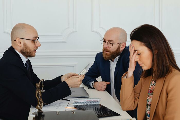 Three people at a table; a male lawyer talks to a couple, the woman looks stressed. Represents lawyers defending impossible cases.