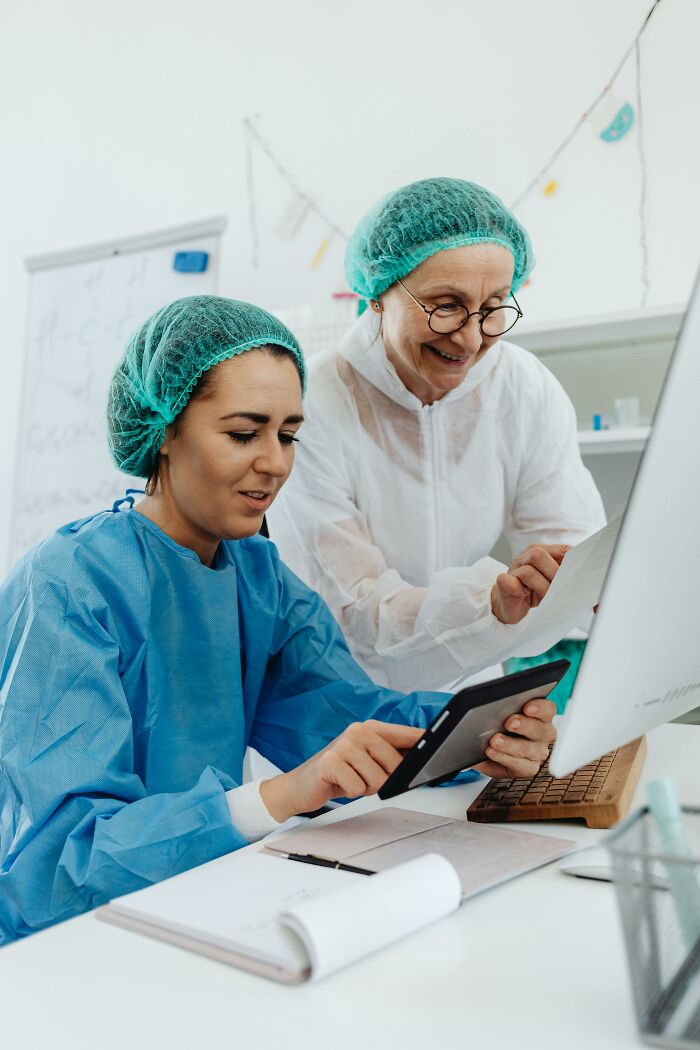 Two medical professionals in scrubs and hairnets reviewing patient charts on a tablet, smiling. Hilarious notes from charts.