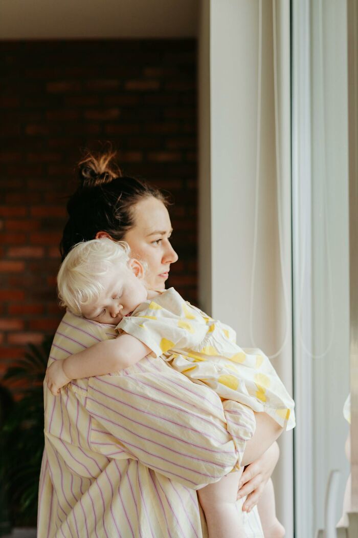 A woman holds a sleeping blonde child, looking out a window. This serene moment reflects on deeply immoral things.