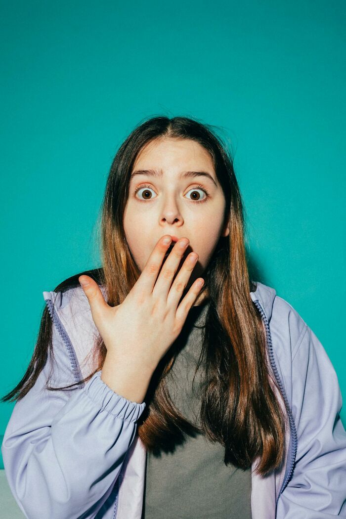 A young person with wide eyes and a hand over their mouth, looking surprised against a teal background, reflecting deeply immoral things.