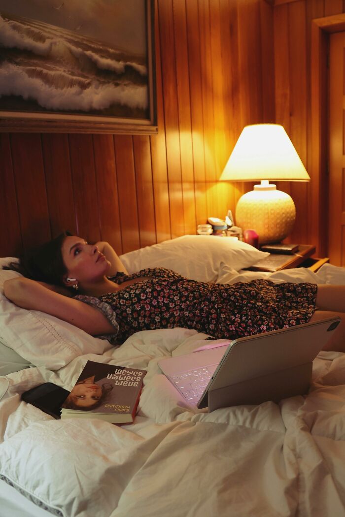 Young woman lying in bed with AirPods, book, and glowing laptop. Gen Z leisure and relaxation at home.