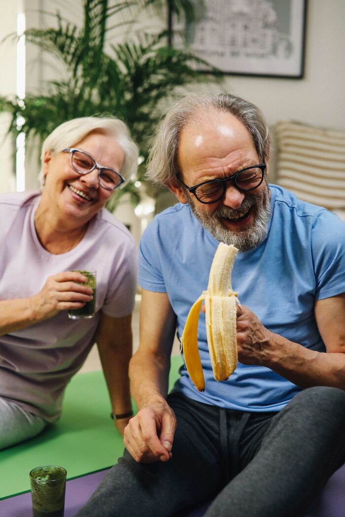 Older couple laughing and eating banana indoors, representing health and happiness in long term partners