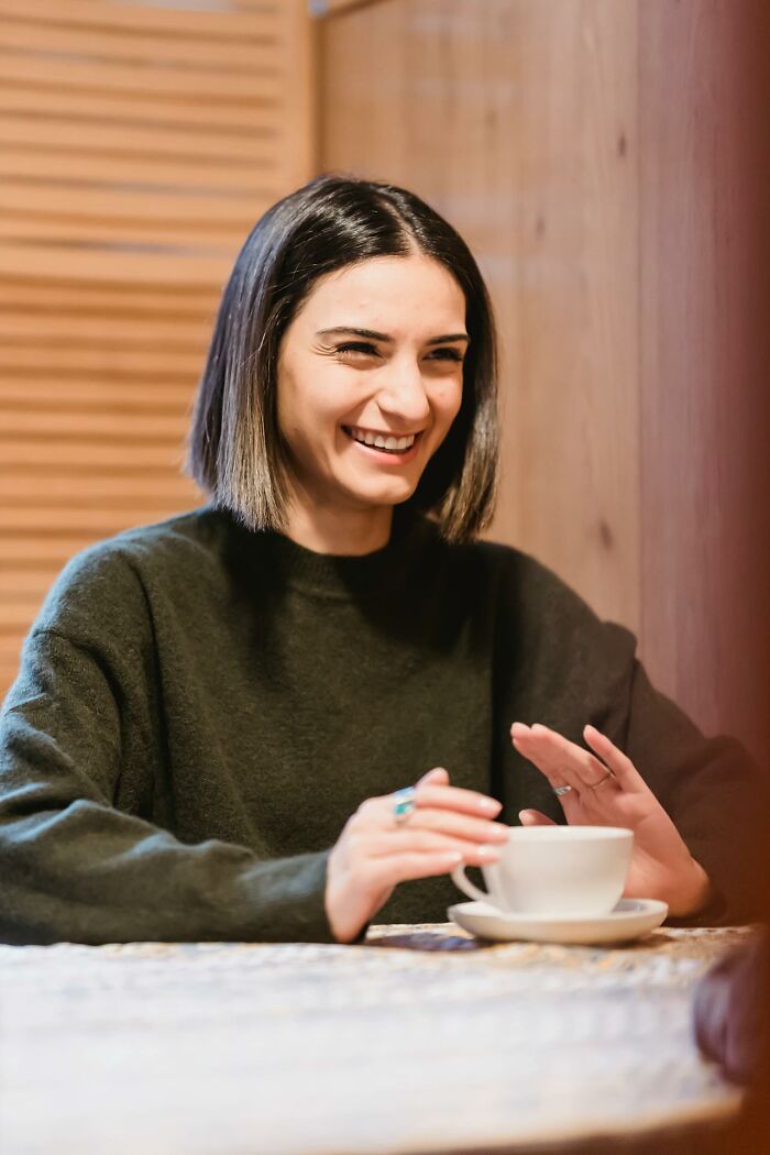 A woman with dark hair laughs, enjoying a drink while listening to wedding drama stories.