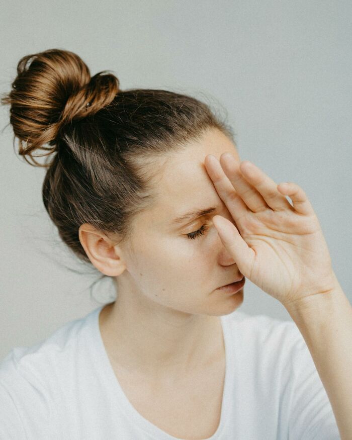 Woman with hand to forehead, eyes closed, appearing stressed or overwhelmed. Reflects dumbfounding moments in the US.