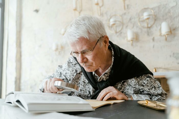 Older man using magnifying glass to read book showing frugal attention to detail