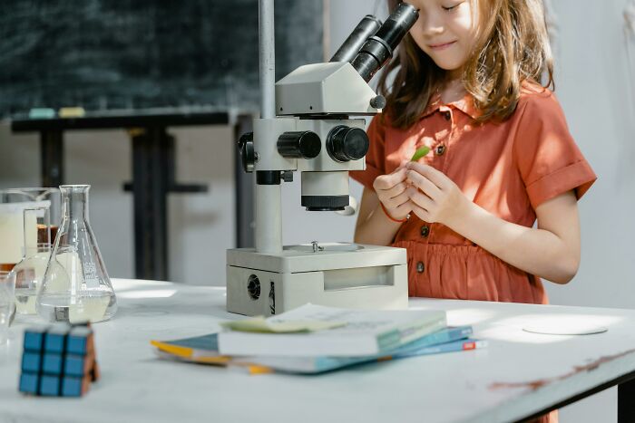 A young girl in an orange dress looking through a microscope, holding a leaf, exploring General Science.