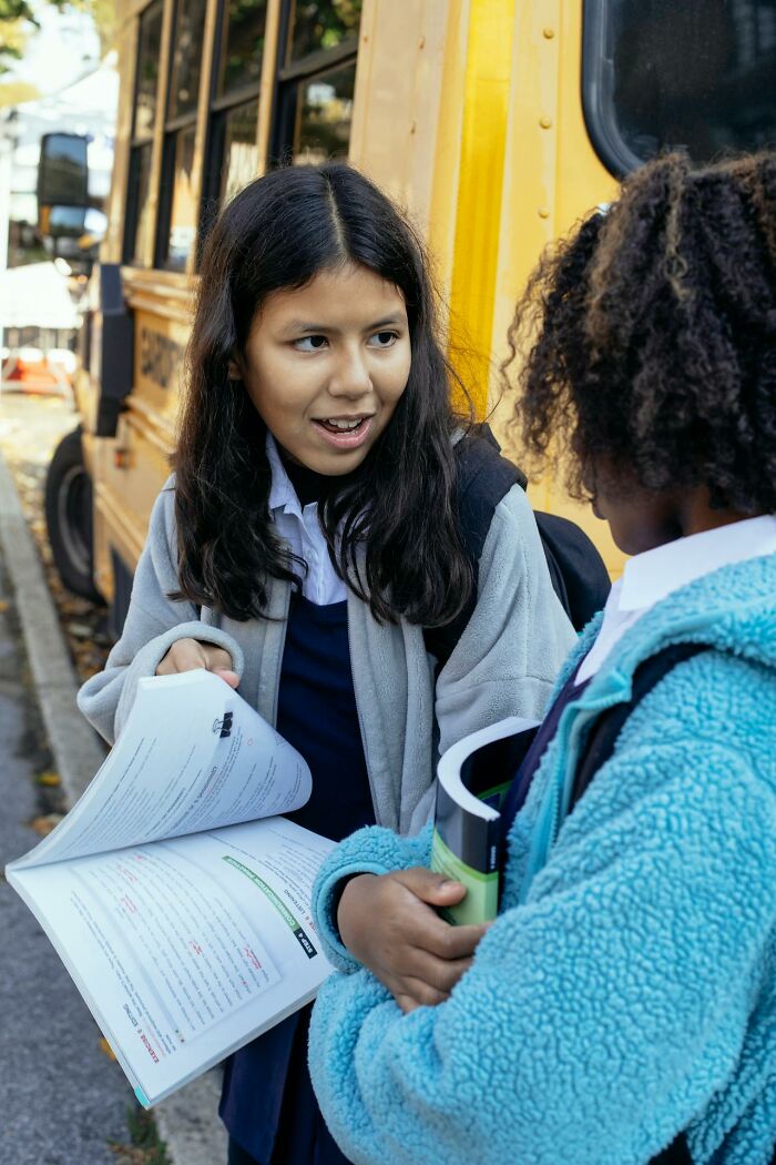 Two teenage girls with backpacks talking near yellow school bus with open book