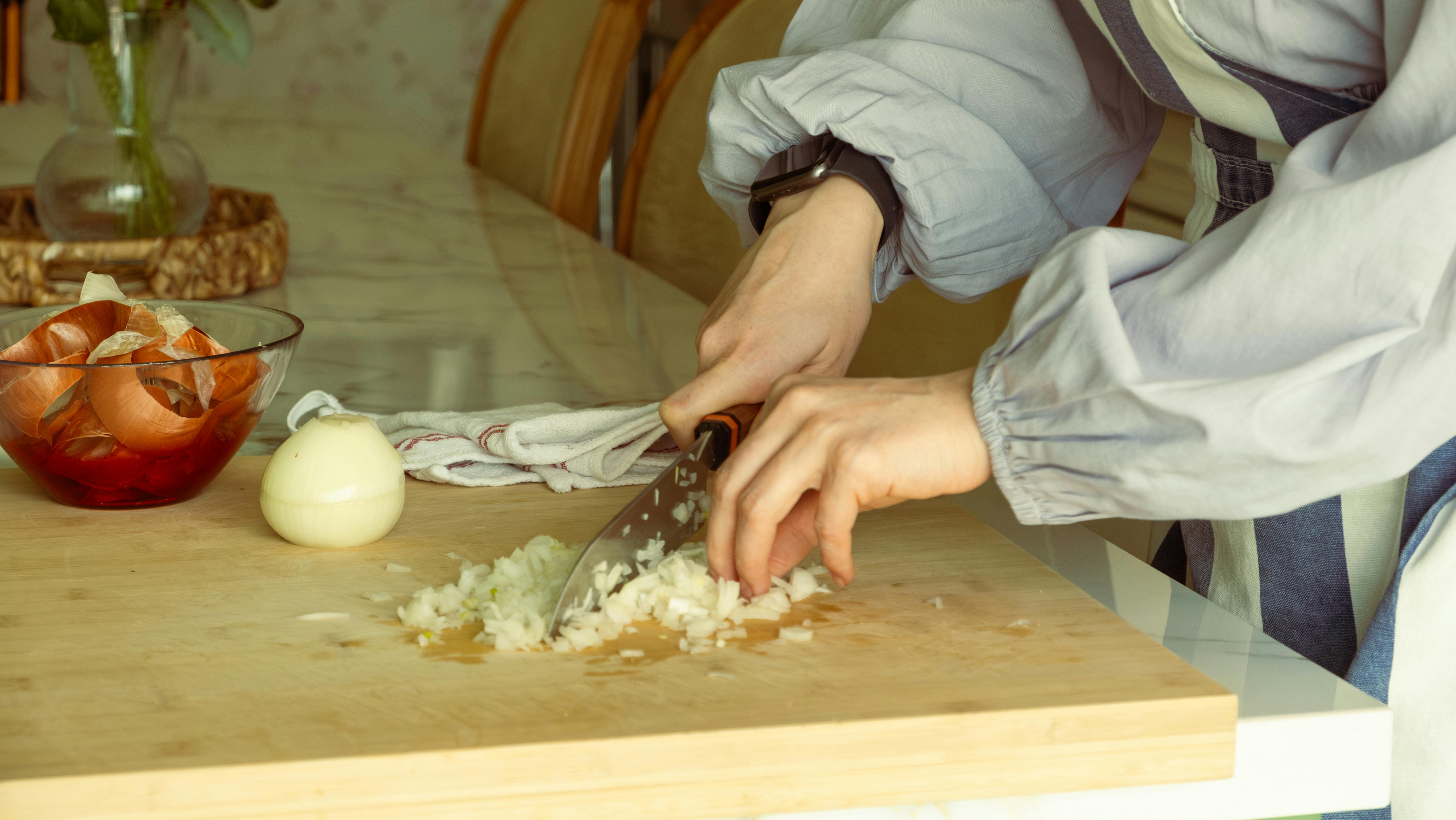 Person chopping onions on a wooden cutting board with onion skins in a bowl nearby