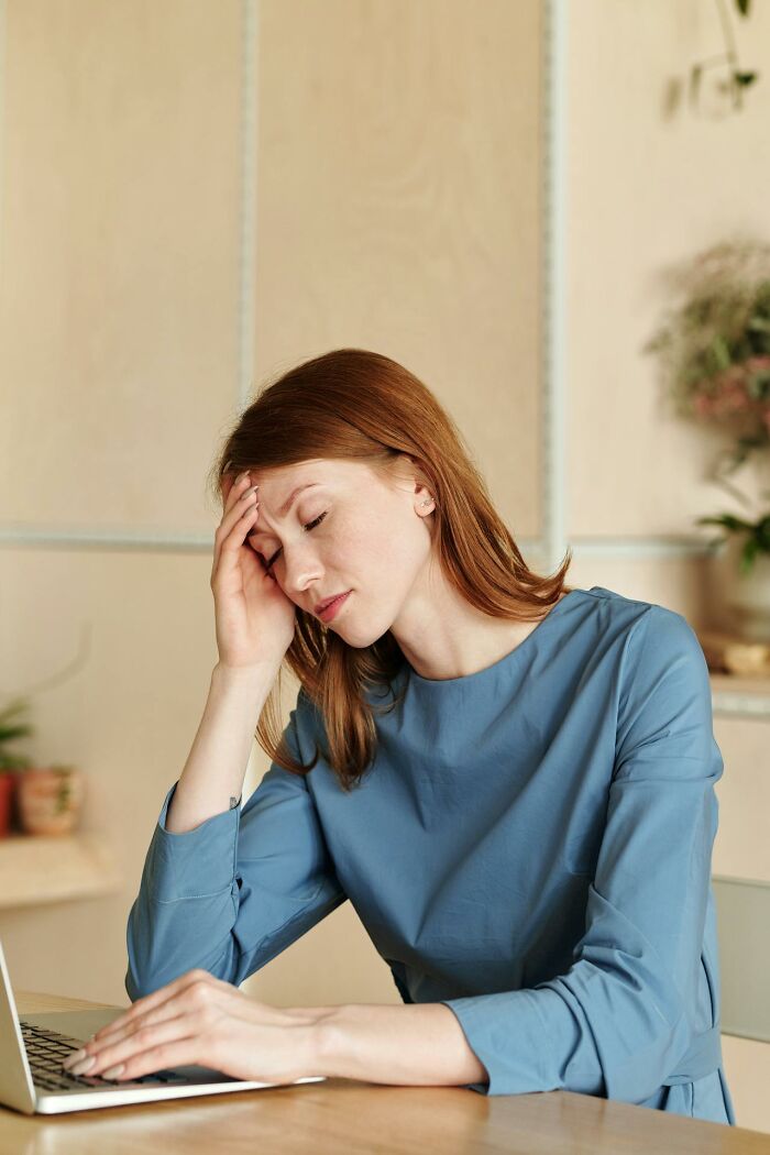 A woman with red hair in a blue top, head in hand, looking stressed while using a laptop. Reflects deeply immoral experiences.