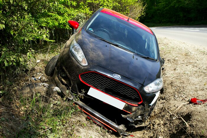 A black car with red trim, crashed into a ditch by the roadside, front bumper broken, revealing a very creepy experience.