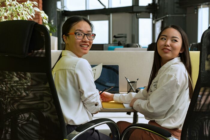Two Gen Z women smiling in an office, one wearing glasses and holding a pencil, capturing generational differences.