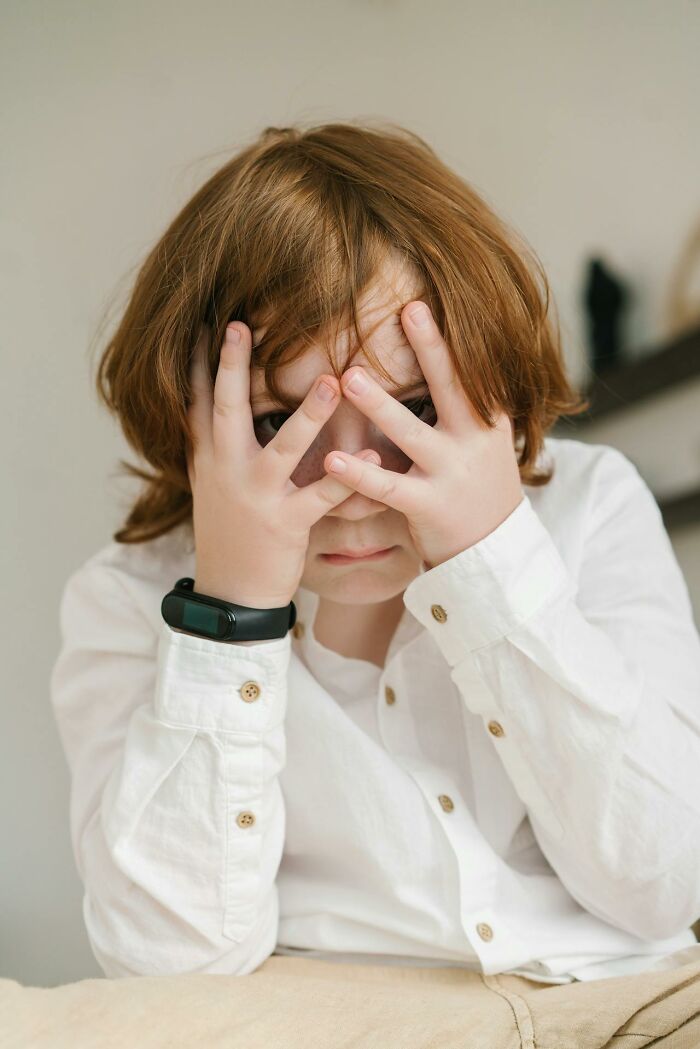 A child with red hair and a white shirt hides their face with hands, peeking through fingers, hinting at deeply immoral things.