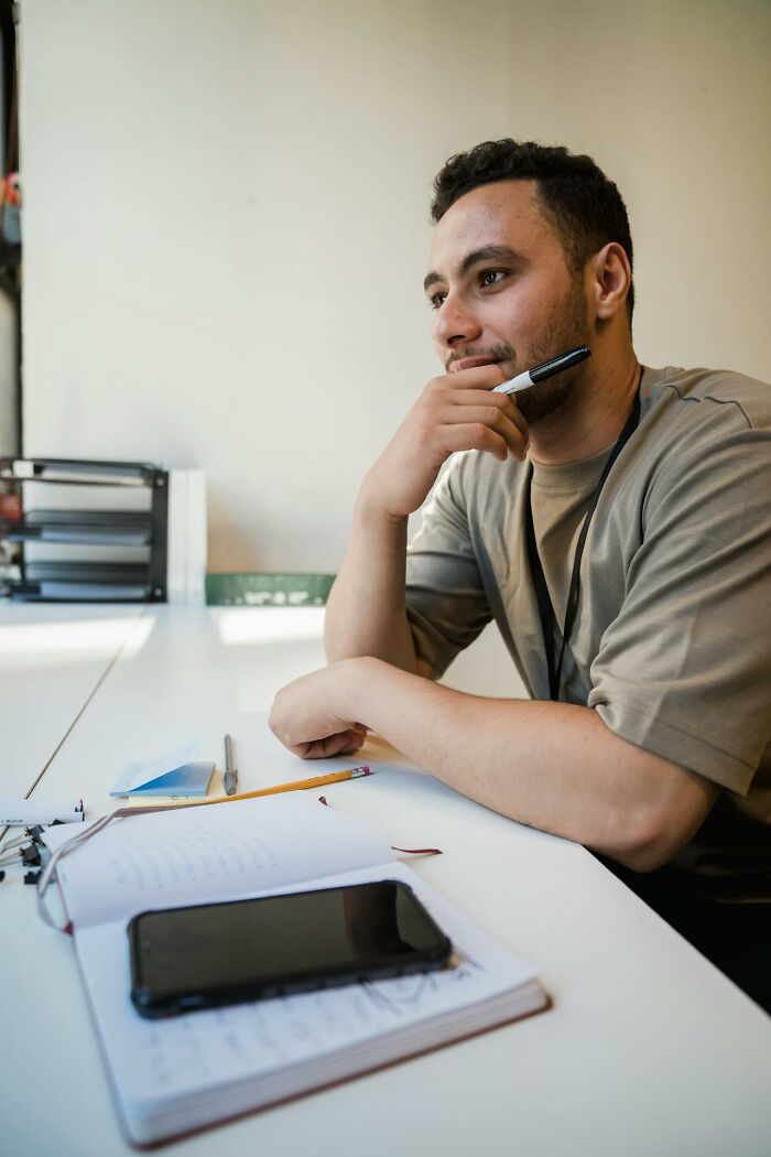 Man thinking with pen near mouth at white desk, laziness saving people idea
