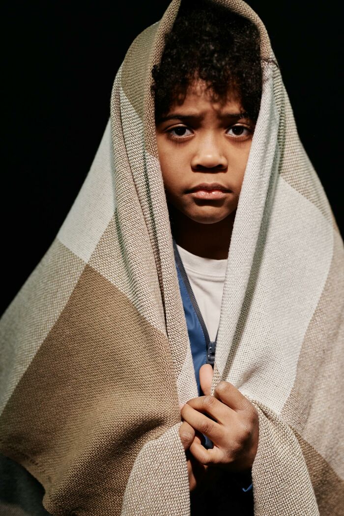 A child with curly hair and a serious expression, wrapped in a checkered blanket. Reflects on deeply immoral things.