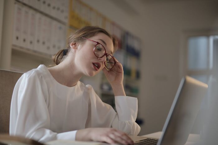 A woman with red-rimmed glasses looking stressed while working on a laptop, depicting potential wedding drama or stress.