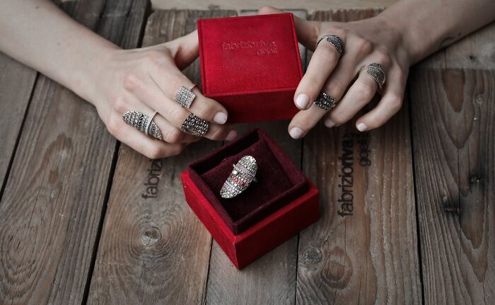 Hands wearing sparkly rings hold a red velvet box. Another ring rests in a red box, questioning reality.