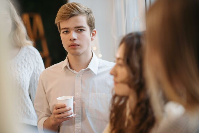 Young Gen Z man in a white shirt holding a cup, looking seriously at the camera, surrounded by blurred people.