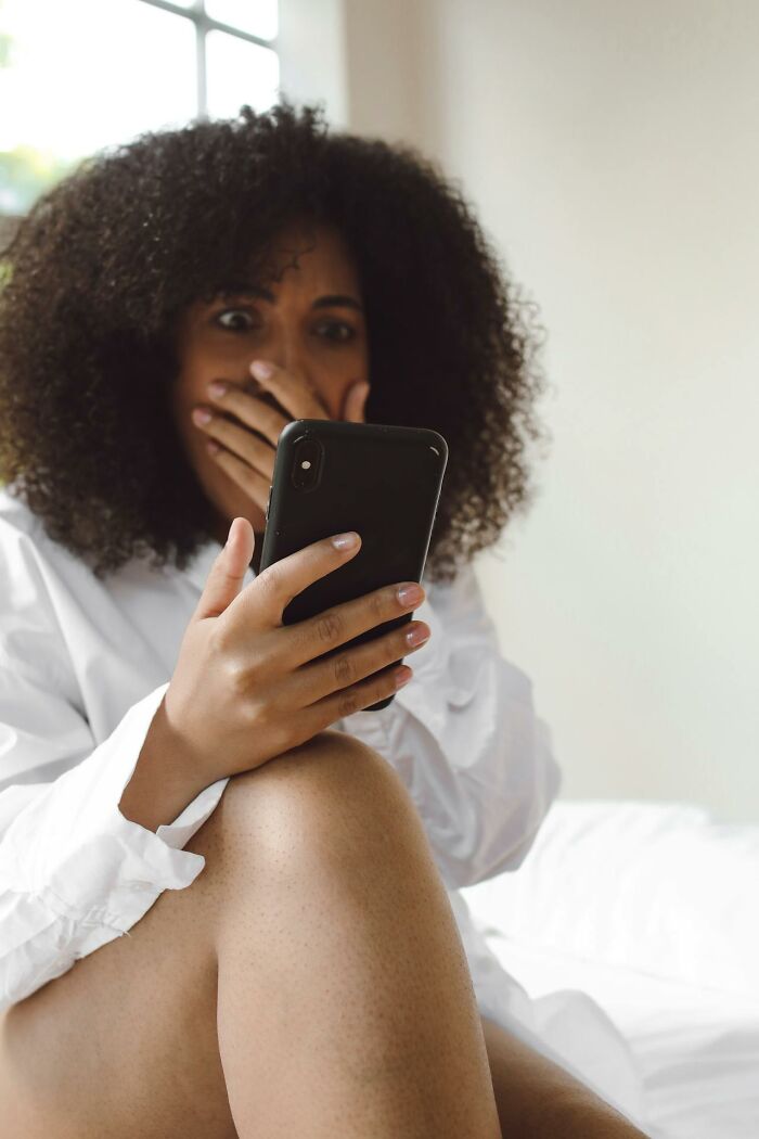A surprised woman with curly hair covers her mouth, holding a phone. She questions reality, finding glitches in the matrix.