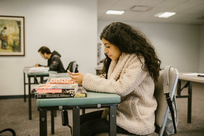 Gen Z student with long dark hair, wearing a cozy coat, distracted by her phone in a classroom. Older generations just don't understand.