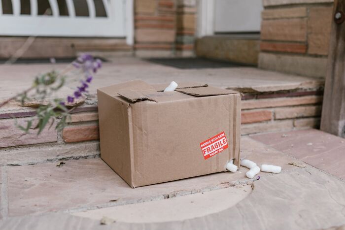 A slightly damaged cardboard box with a red FRAGILE sticker and packing peanuts spilling out, on stone steps. Engagement stories often involve unexpected chaos!
