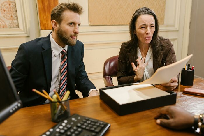 Two lawyers, a man and a woman, discuss difficult cases. The woman holds a document, gesturing, while the man listens intently.