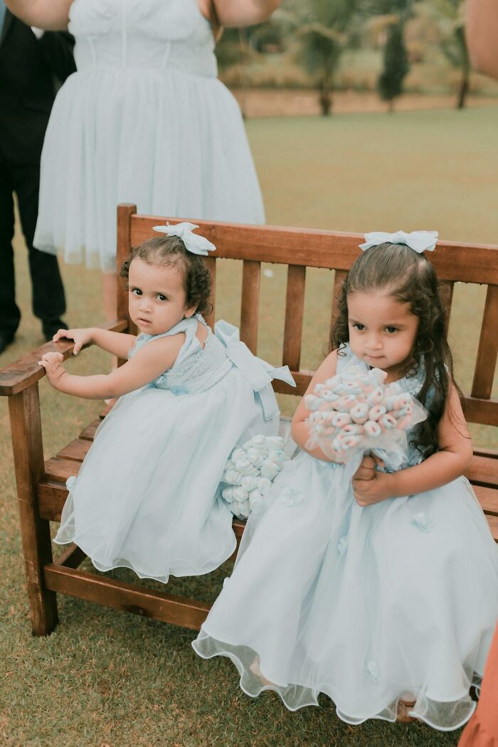 Two young flower girls in light blue dresses sitting on a wooden bench, holding small bouquets at a wedding event.