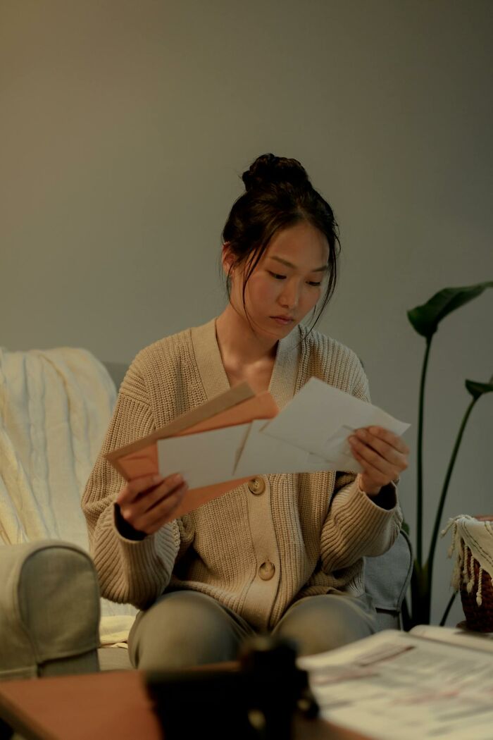 A young woman with a bun, wearing a beige cardigan, looks thoughtfully at envelopes. She's processing something deeply immoral.