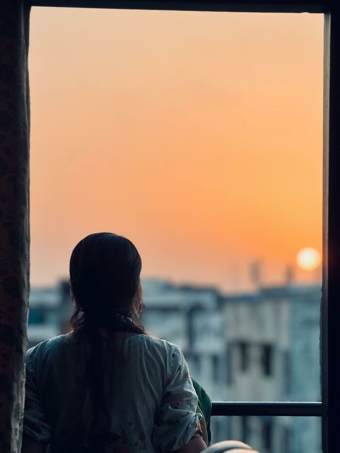Woman standing on balcony looking at orange sunset over cityscape