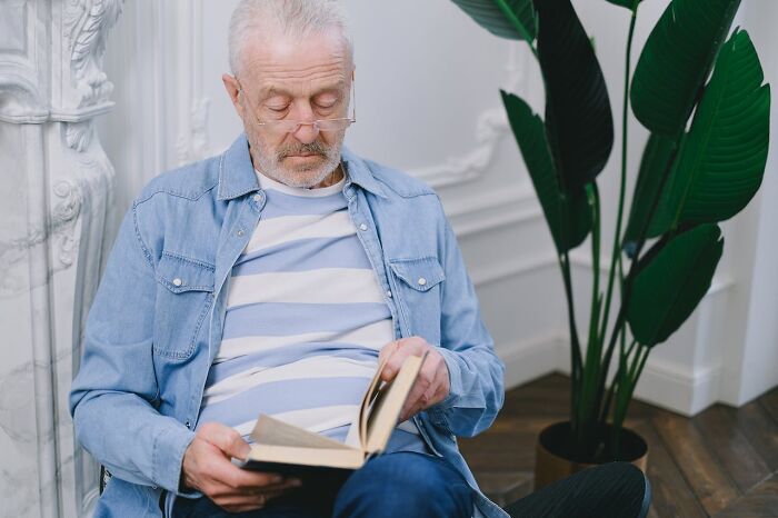 A senior man with glasses and a blue striped shirt reads a book. He questions reality, perhaps experiencing glitches in the matrix.