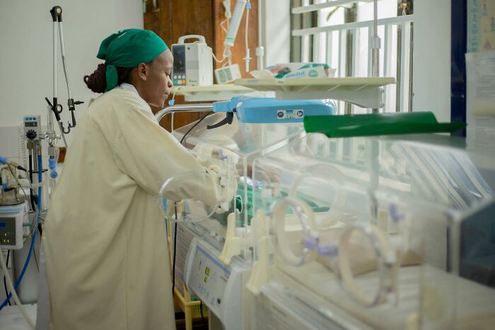 A medical professional in a green scrub cap tending to a baby in an incubator, surrounded by medical charts.